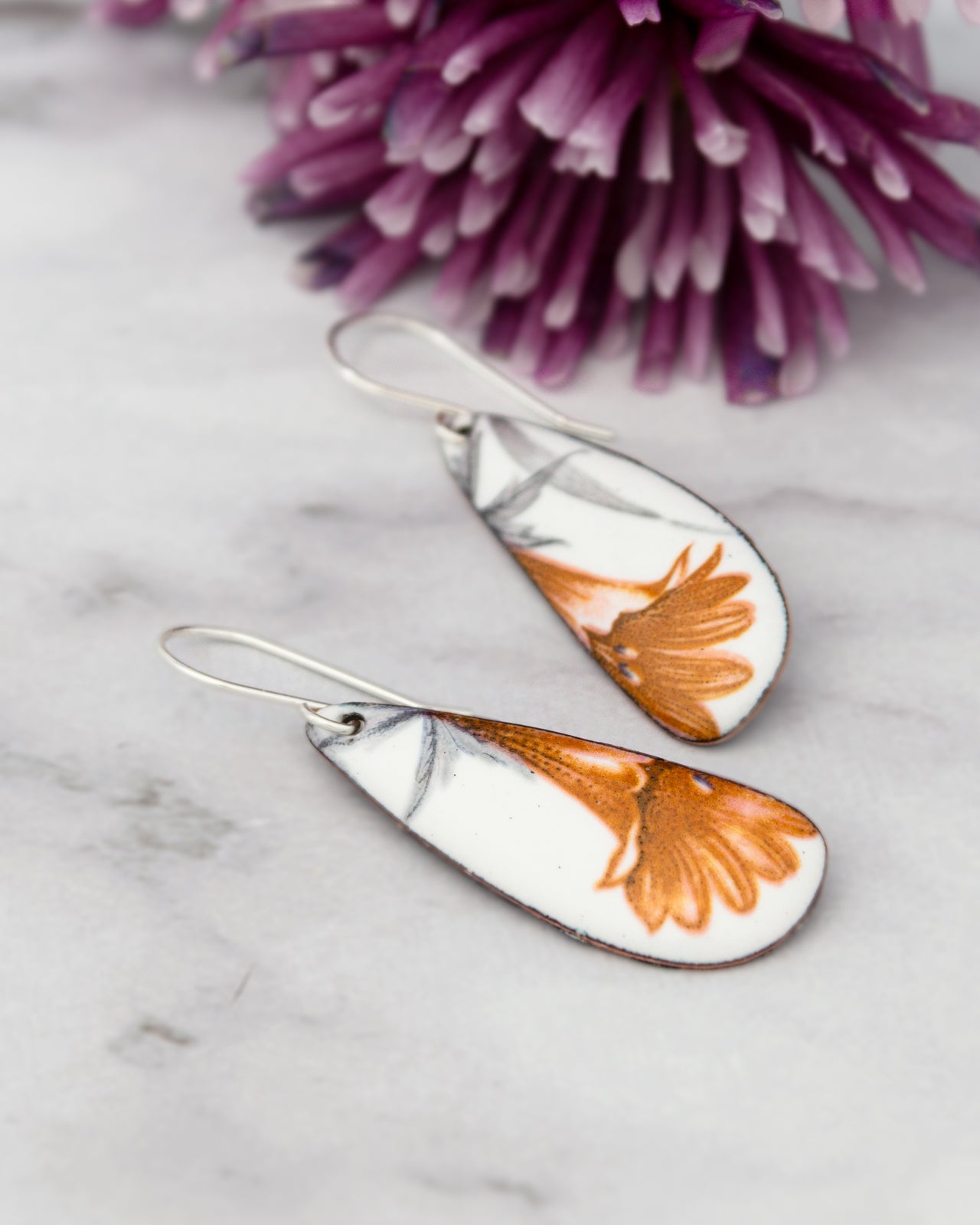 A pair of earrings with a floral design, featuring orange and white colors, are placed on a marble surface next to a purple flower.