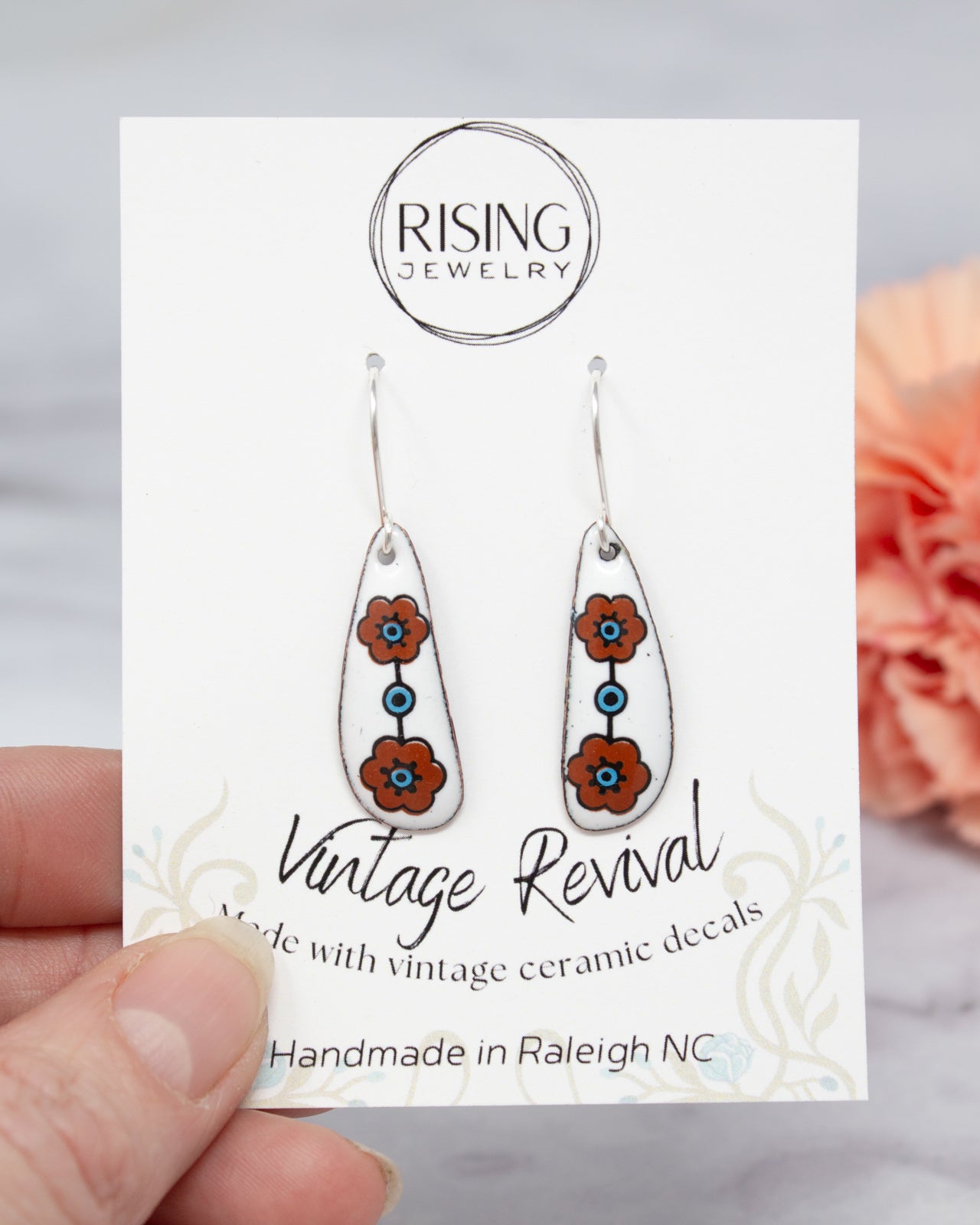 A hand holding a pair of earrings with a white card displaying the earrings and the company's name.