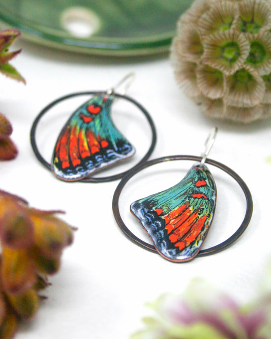 a pair of colorful butterfly-shaped earrings with intricate designs, placed on a white surface alongside some flowers.