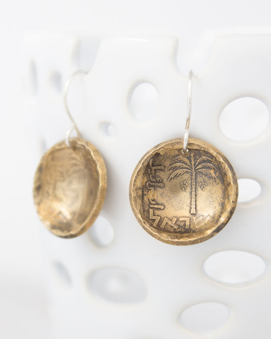 two round, metallic earrings hanging on a white surface. The earrings have a design that includes a palm tree and some text or symbols.