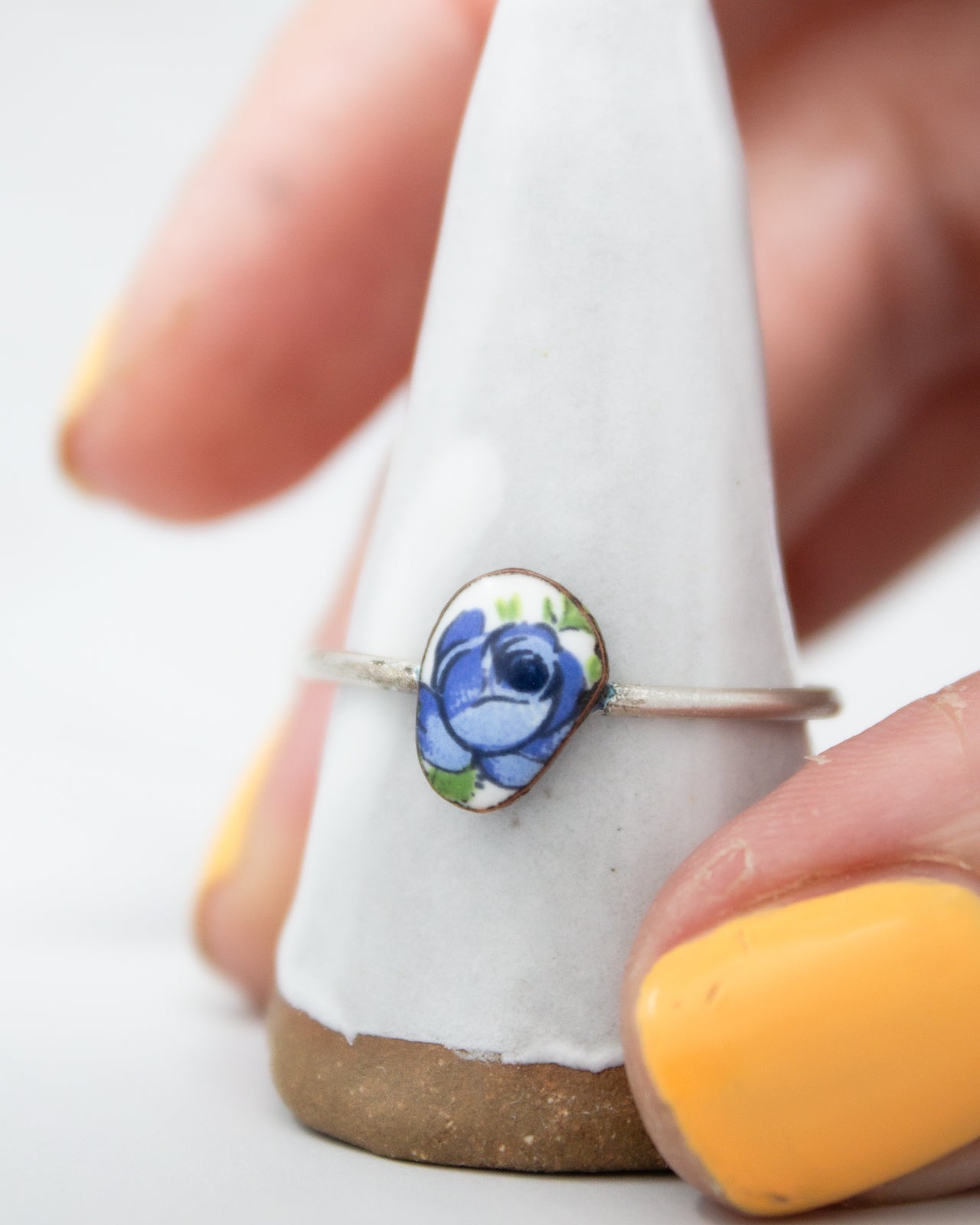 A hand is holding a white ring with a blue flower design on it, placed on a white stand.