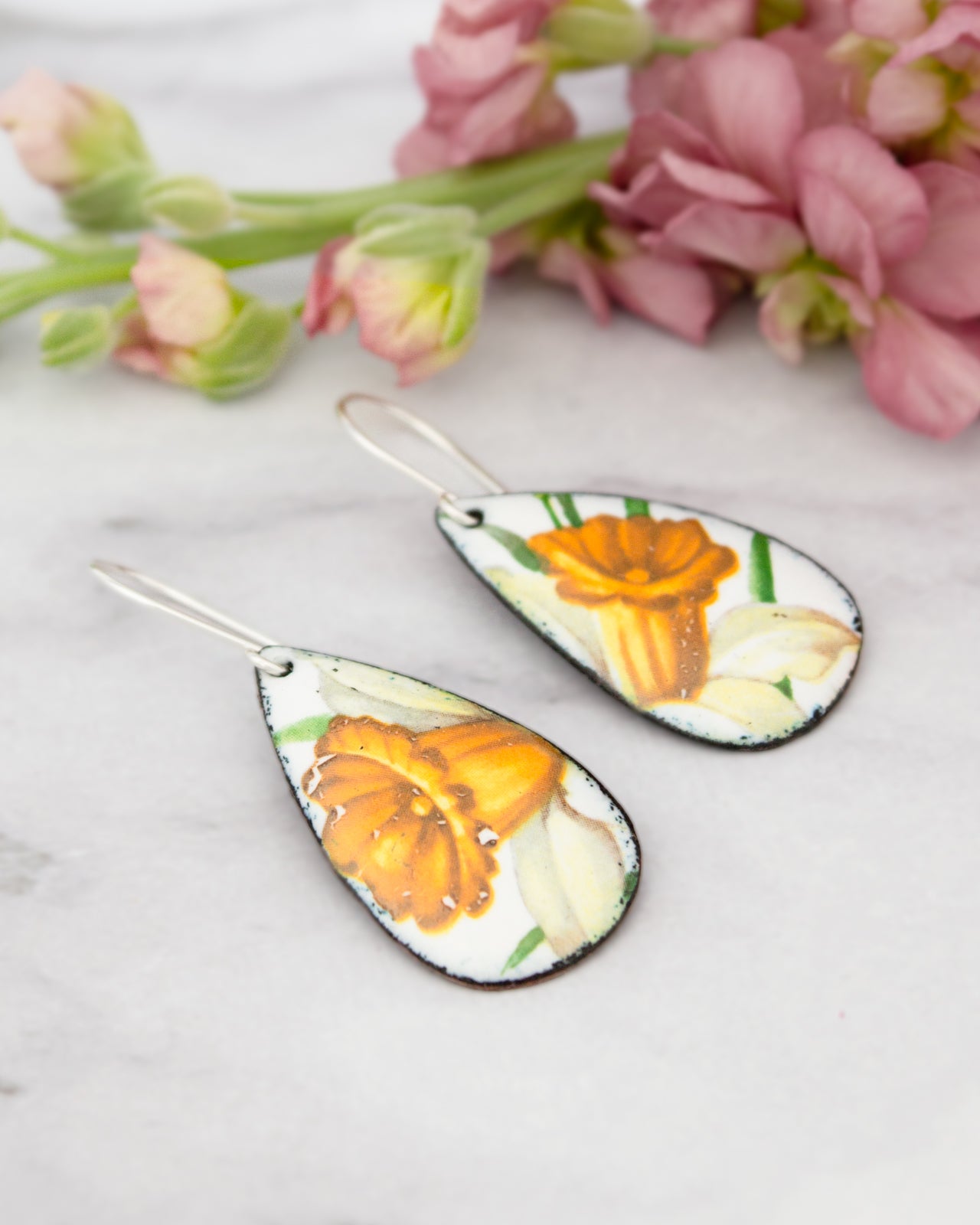 A pair of earrings with a floral design, featuring yellow and orange flowers, are displayed on a white surface alongside a bouquet of pink flowers.