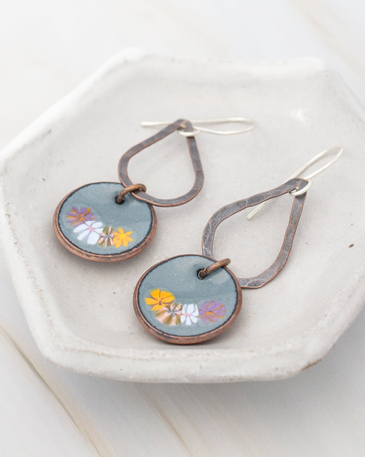 A pair of earrings with a floral design on the circular pendants, resting on a white plate.