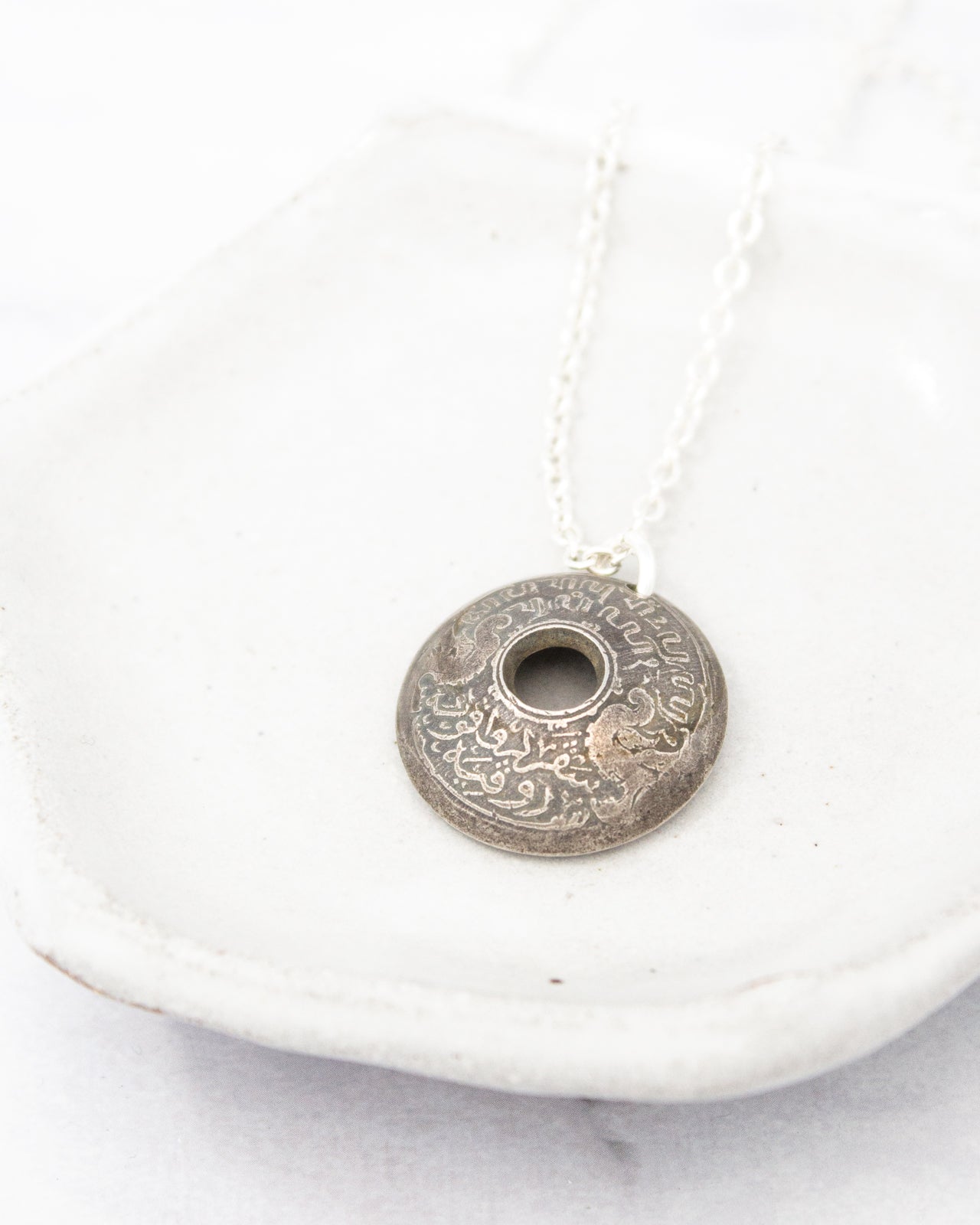 A silver pendant with an intricate design is displayed on a white plate.