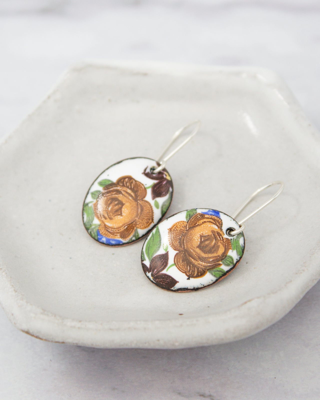 A pair of oval-shaped earrings with a floral design, resting on a white plate or tray.