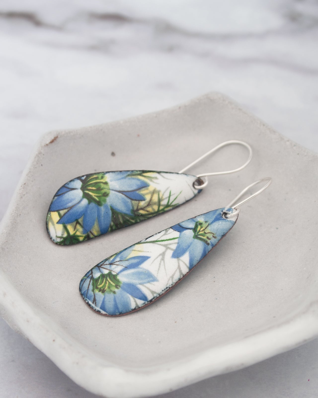 A pair of earrings with blue and green floral designs, resting on a white plate or bowl.