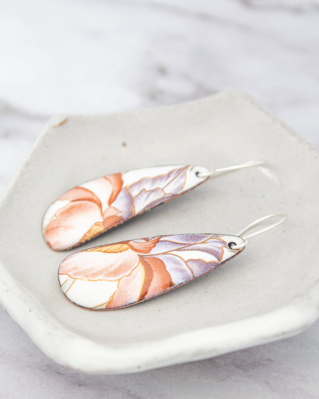 A pair of earrings with a floral pattern on the top half and a metallic finish on the bottom half, resting on a white plate against a marble background.