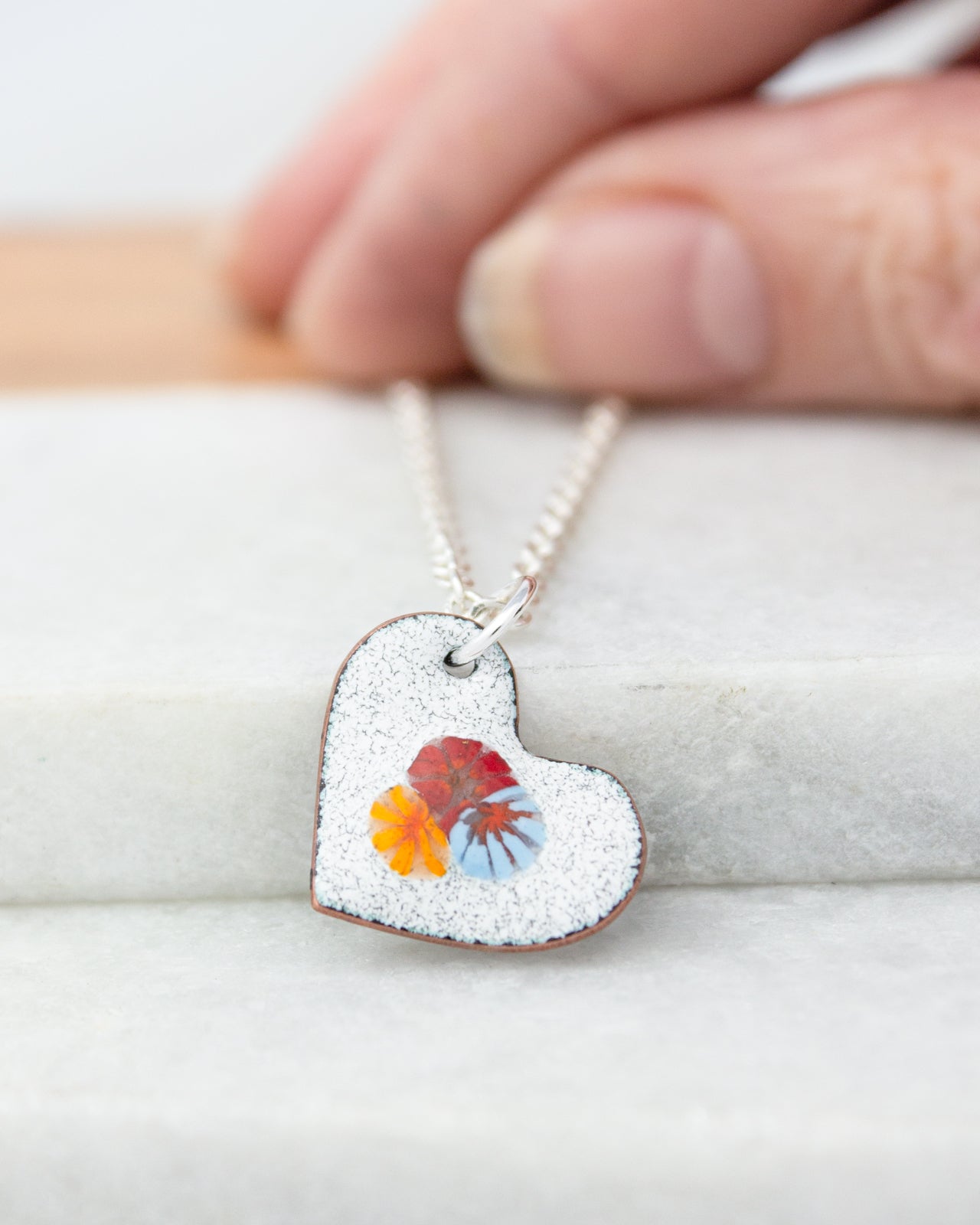 A hand is holding a heart-shaped pendant with a floral design, resting on a white surface.