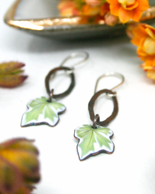 a pair of earrings with green leaf-shaped pendants, placed on a white surface alongside some orange flowers.