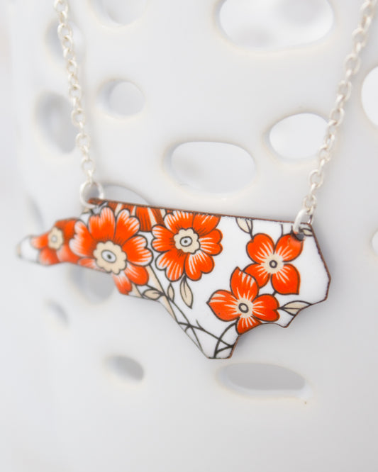 A necklace with a pendant in the shape of the state of North Carolina, featuring a floral design in orange and white colors.