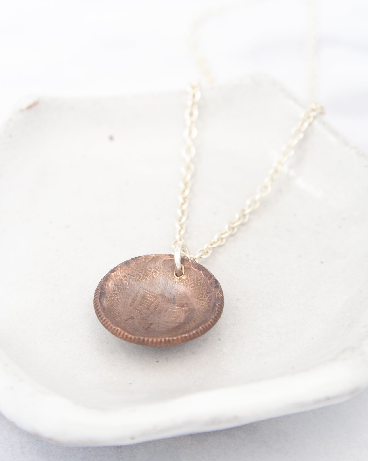 A necklace with a round pendant made of copper-colored metal, possibly a coin, is displayed on a white surface.