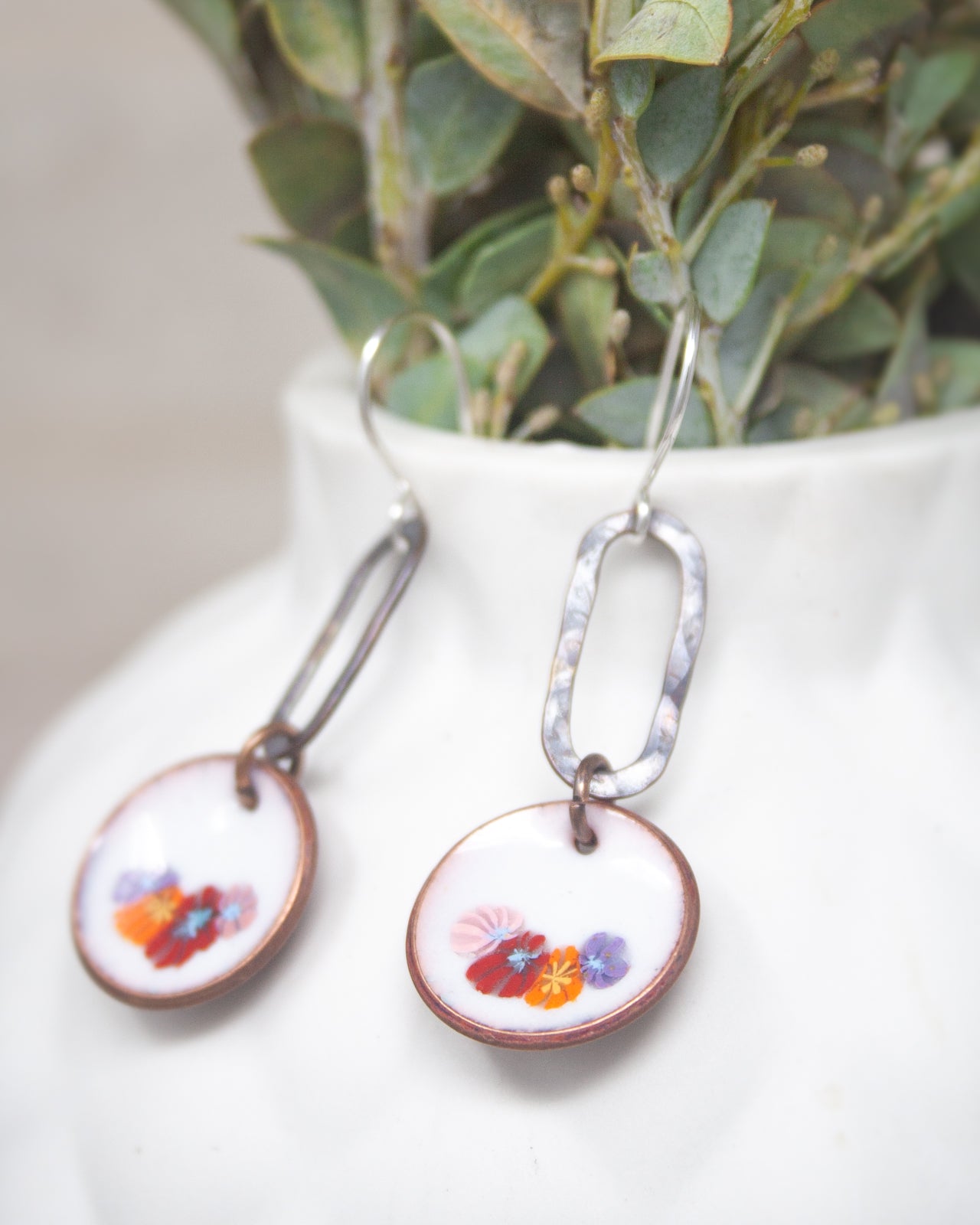 A pair of earrings with colorful floral designs, hanging from a white vase with green leaves.