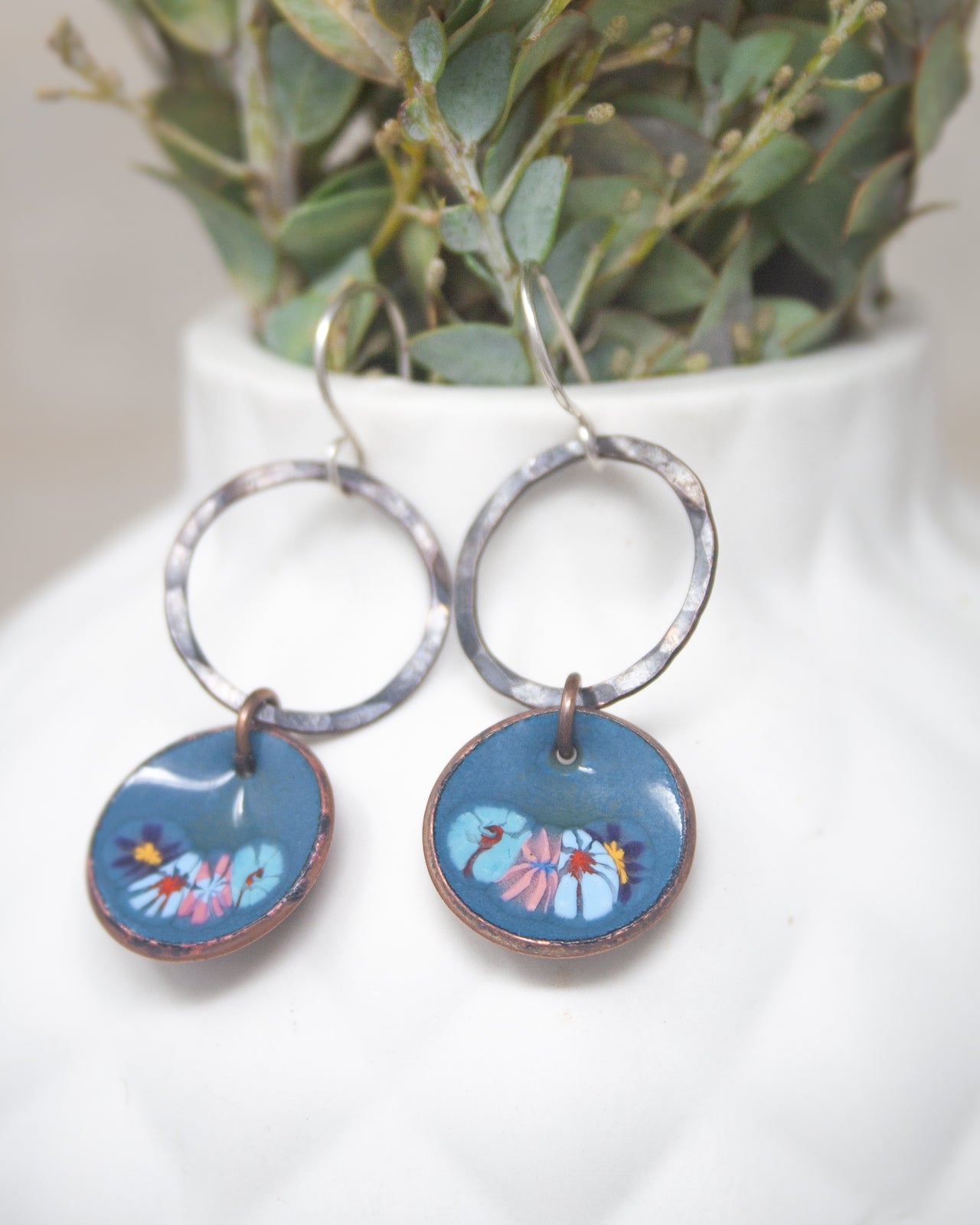 A pair of blue earrings with circular designs featuring flowers, hanging from a white vase with greenery.