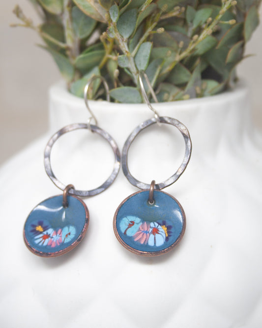A pair of blue earrings with circular designs featuring flowers, hanging from a white vase with greenery.