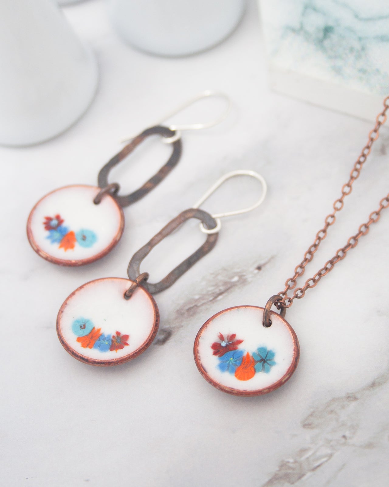 a necklace and earrings with floral designs, including blue, orange, and red flowers, displayed on a marble surface.