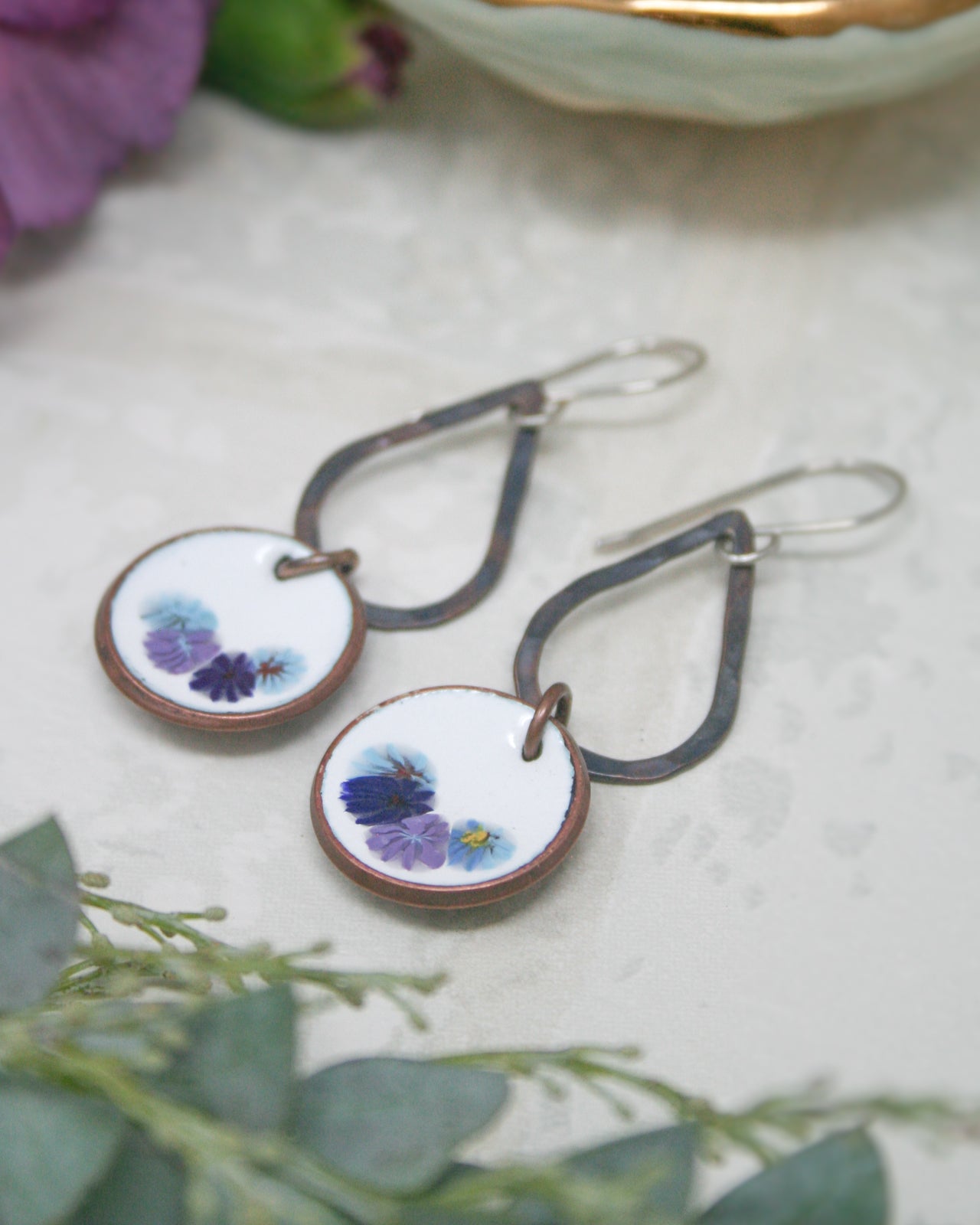 A pair of earrings with floral designs on the circular pendants, placed on a white surface with some greenery in the background.