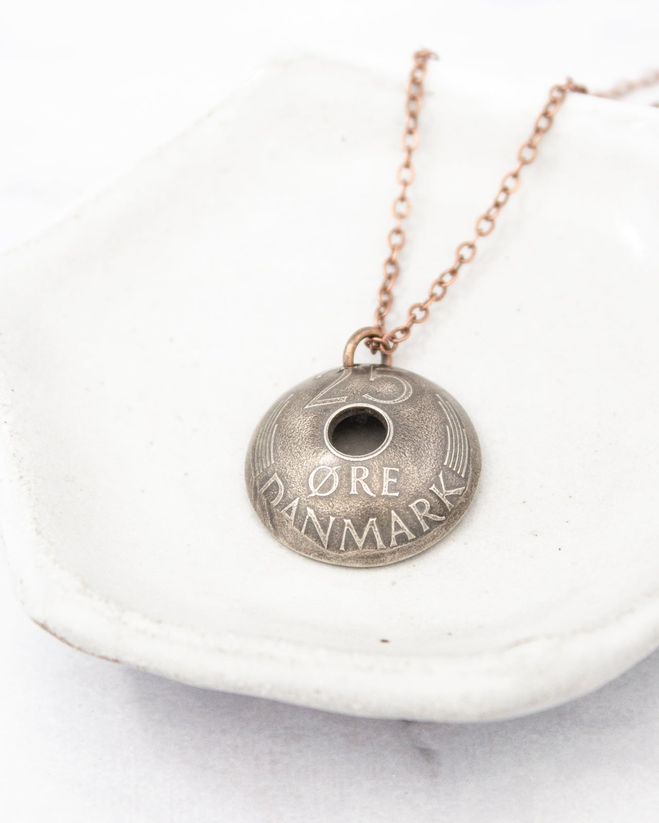 A silver pendant necklace with the text "DANMARK" engraved on it, resting on a white surface.