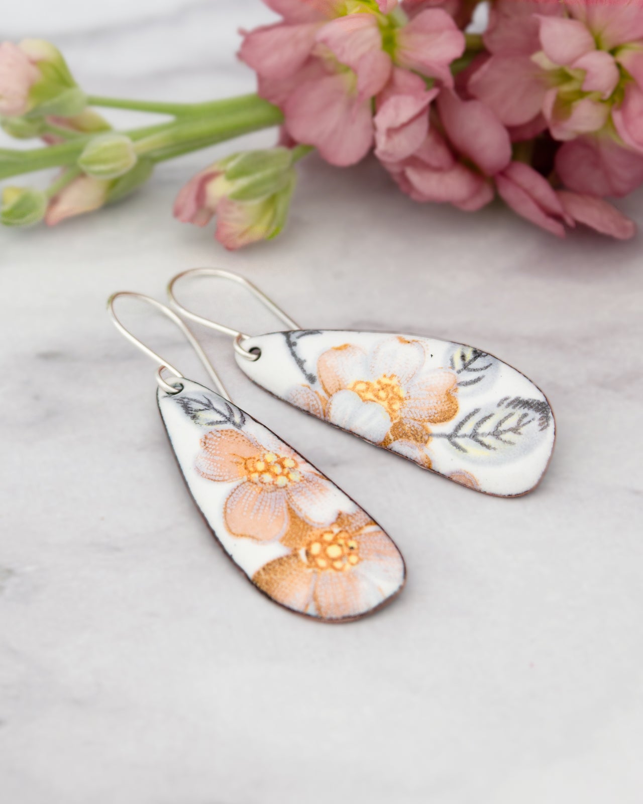 A pair of white earrings with a floral design, placed on a marble surface next to a bouquet of pink flowers.