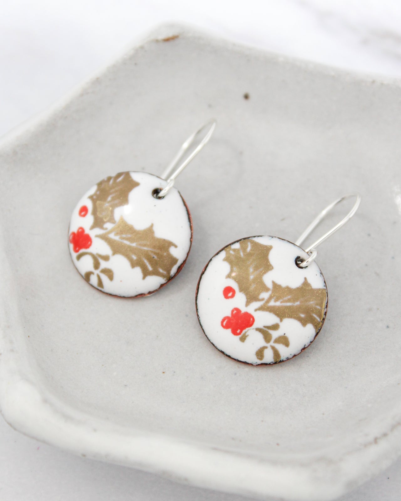 A pair of round earrings with a white and gold floral design, featuring red berries and leaves, resting on a white plate.
