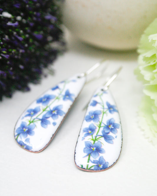 A pair of white earrings with blue flower designs on them, placed on a white surface next to some purple flowers.