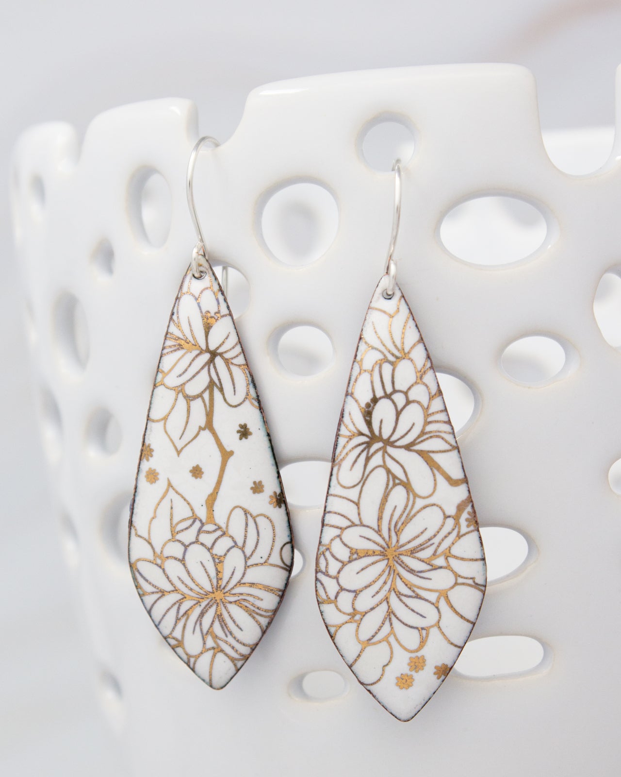 A pair of white earrings with gold floral designs, hanging from a white perforated holder.