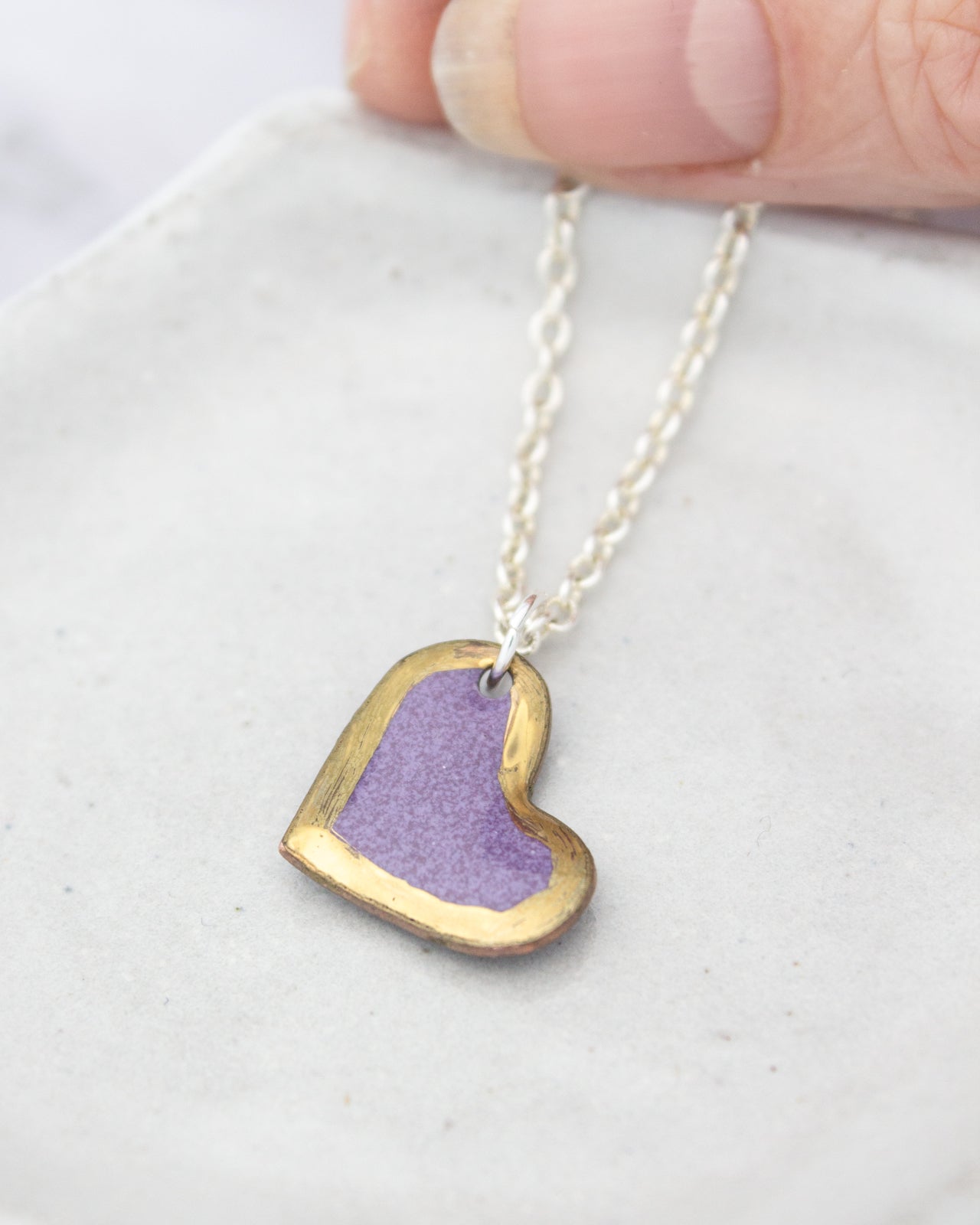 A hand is holding a delicate necklace with a heart-shaped pendant in a vibrant purple color.
