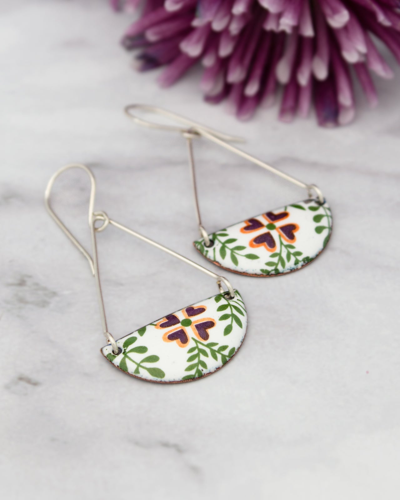 A pair of earrings with a floral design on a white marble surface, with a purple flower in the background.