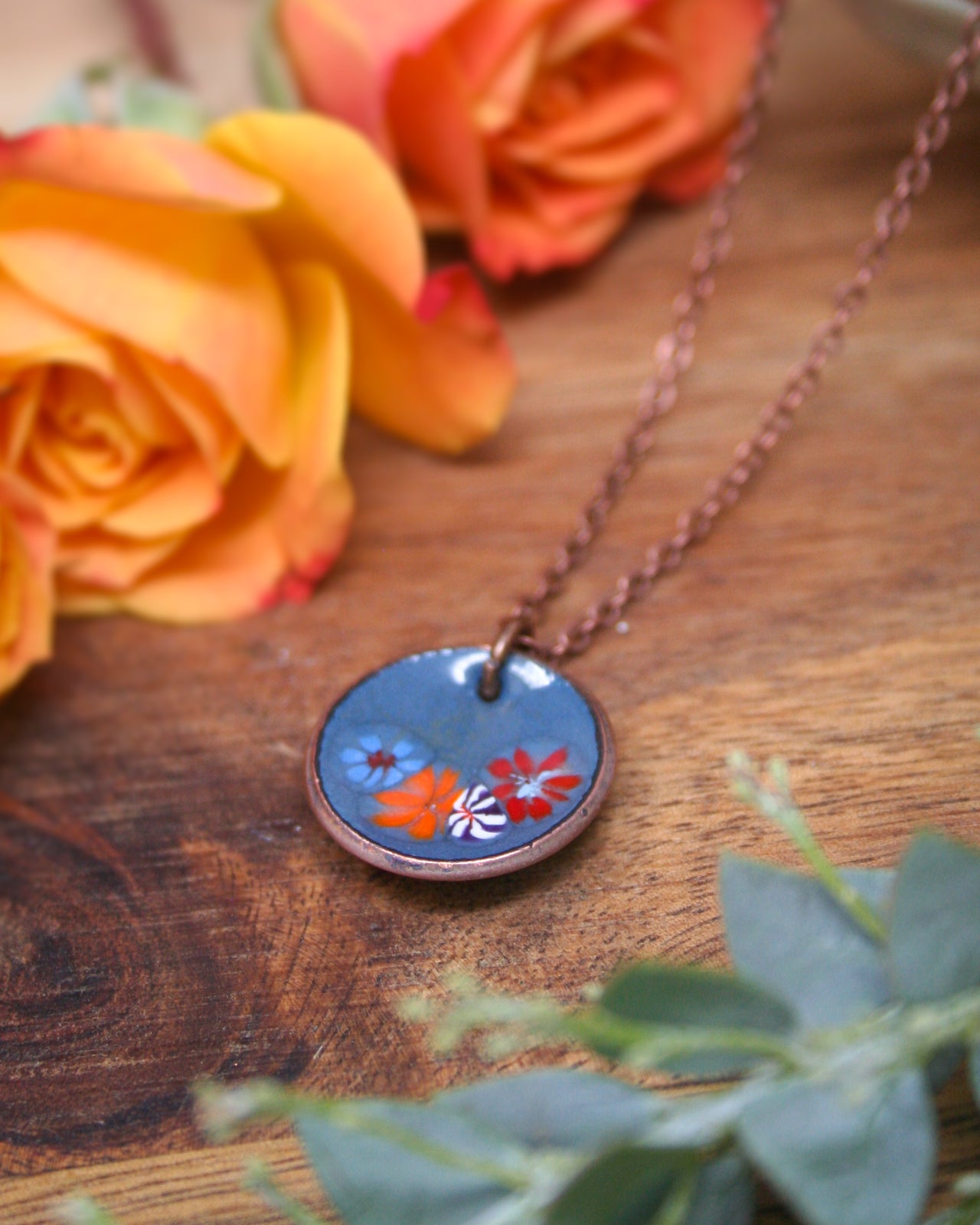 A blue pendant necklace with a floral design rests on a wooden surface, surrounded by vibrant orange and yellow roses.