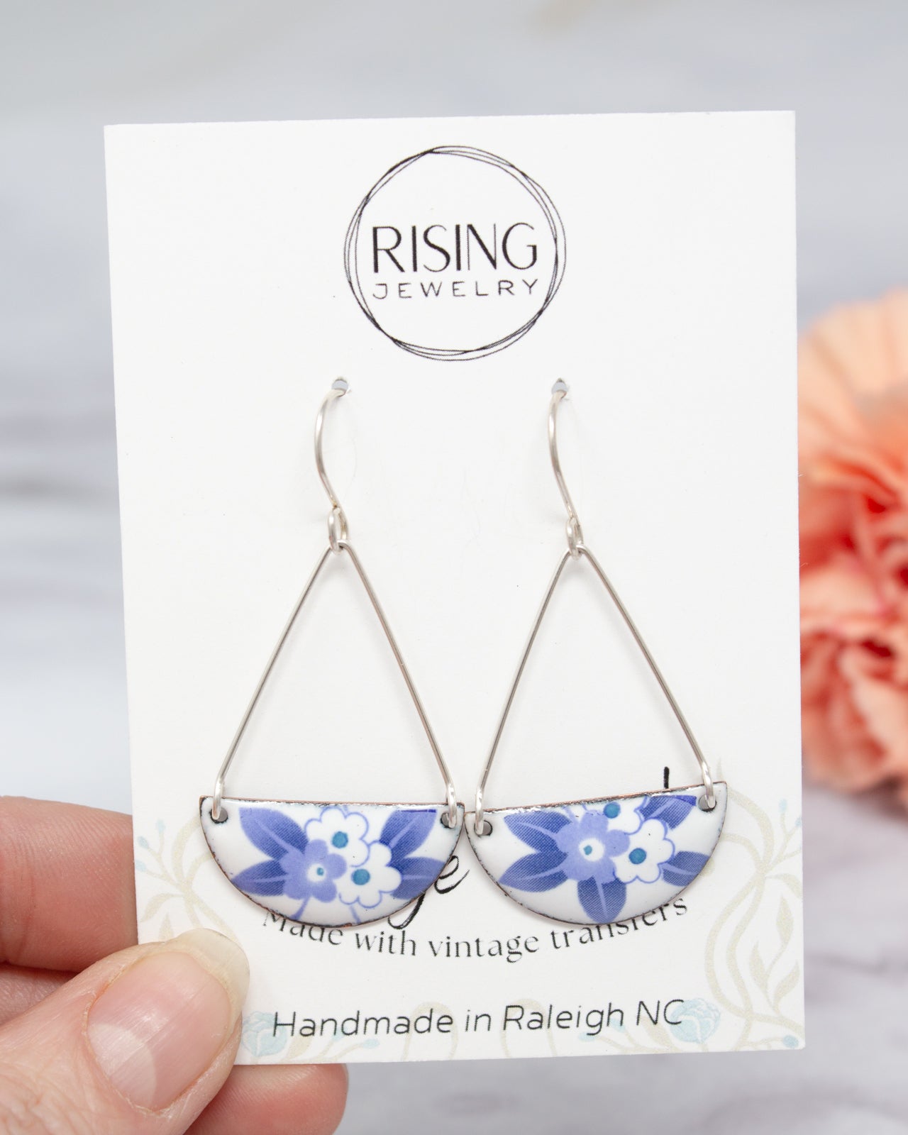 A hand holding a pair of earrings with a floral design, against a blurred background.