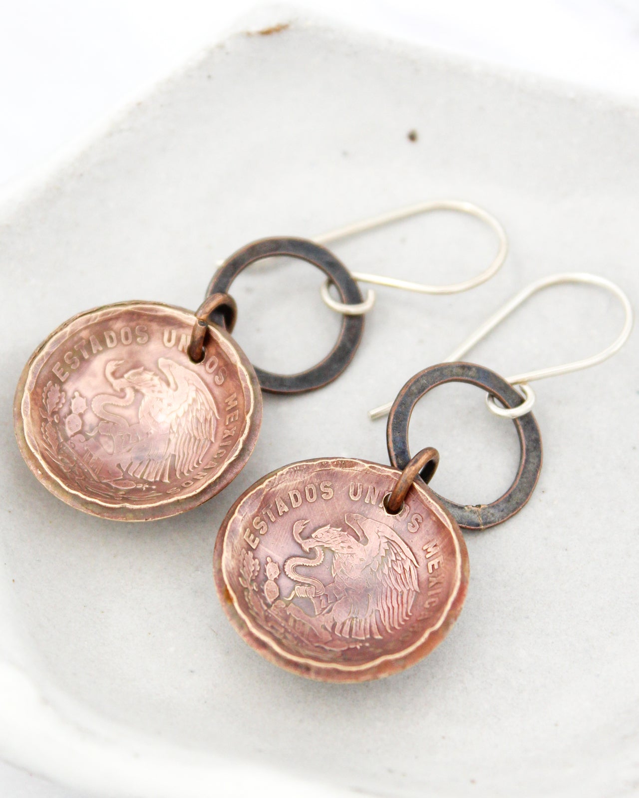 A pair of earrings made from two different colored metal pieces, with one piece being copper and the other being silver. The earrings have a circular design with a textured surface and a metal ring attached to the top.