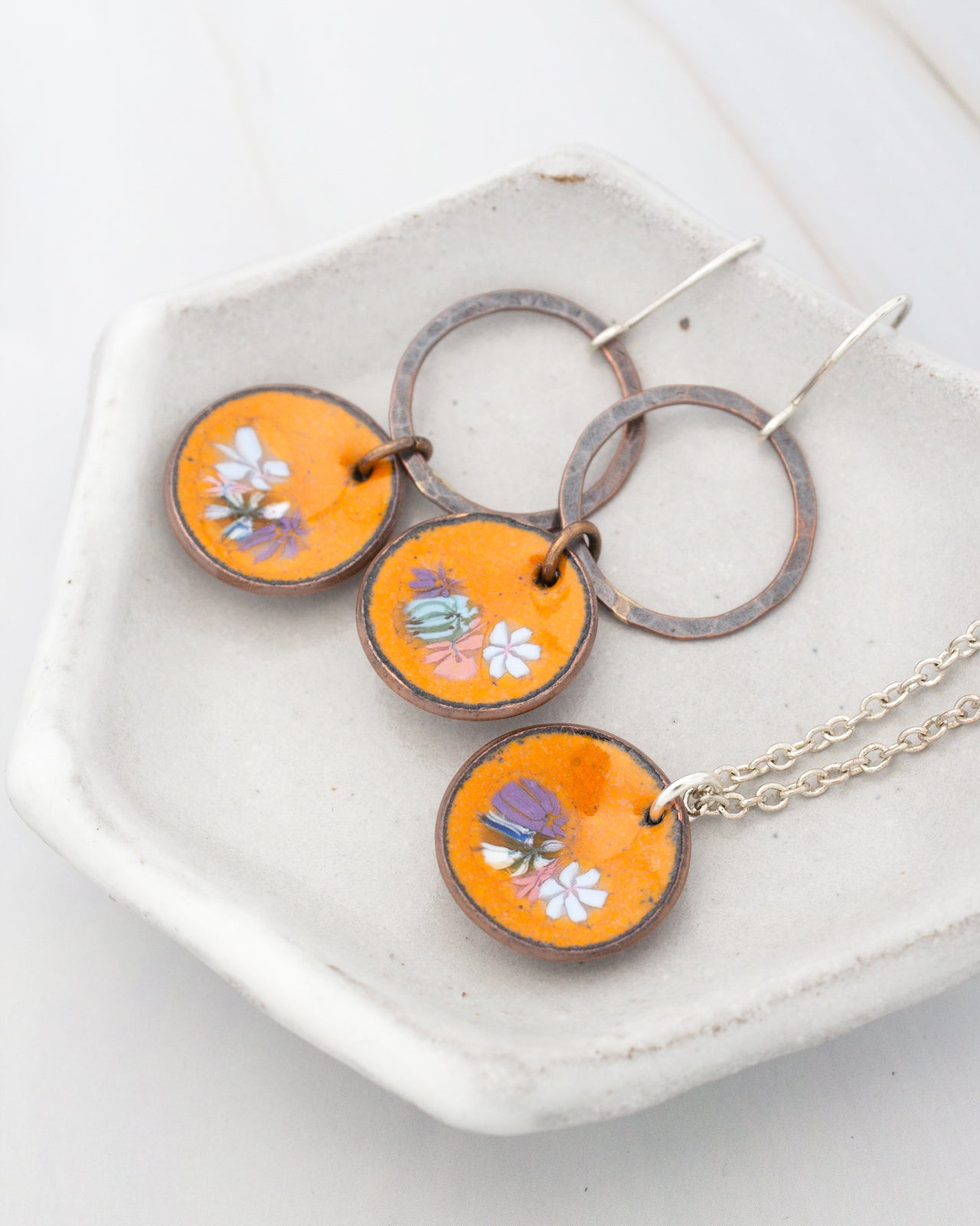 A pair of orange enamel earrings with floral designs and a necklace with matching pendants are displayed on a white plate.