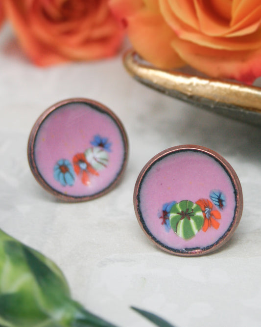 two round, colorful earrings with floral designs on a white surface, with a blurred background of orange and pink flowers.