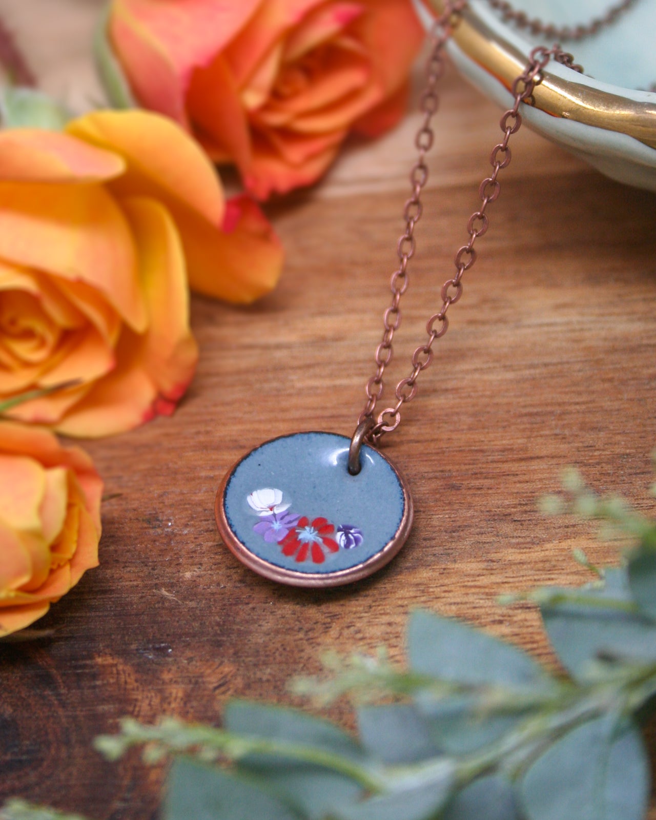 a necklace with a blue circular pendant adorned with a floral design, surrounded by vibrant orange and yellow roses on a wooden surface.