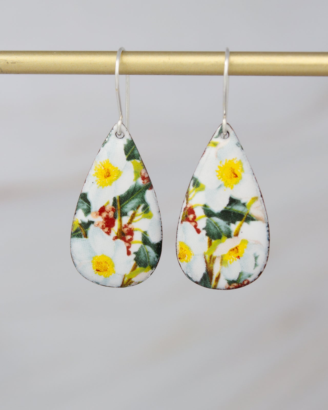 A pair of earrings with a floral pattern, featuring yellow flowers and green leaves, hanging from a gold-colored metal bar.