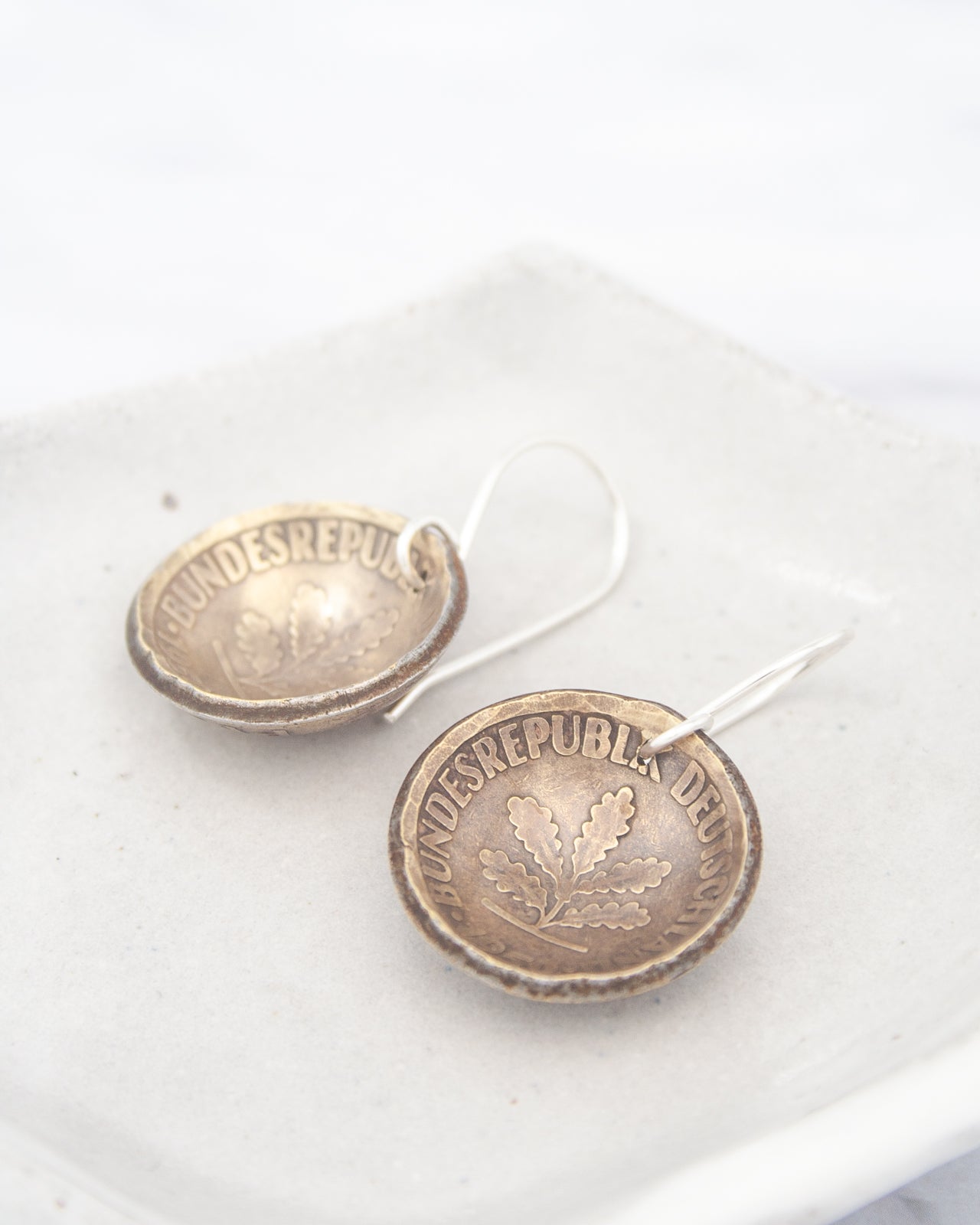 a pair of earrings made of a shiny metal, possibly silver, with the word "BUNDESREPULS" engraved on them. The earrings are placed on a white surface, such as a plate or a tray.