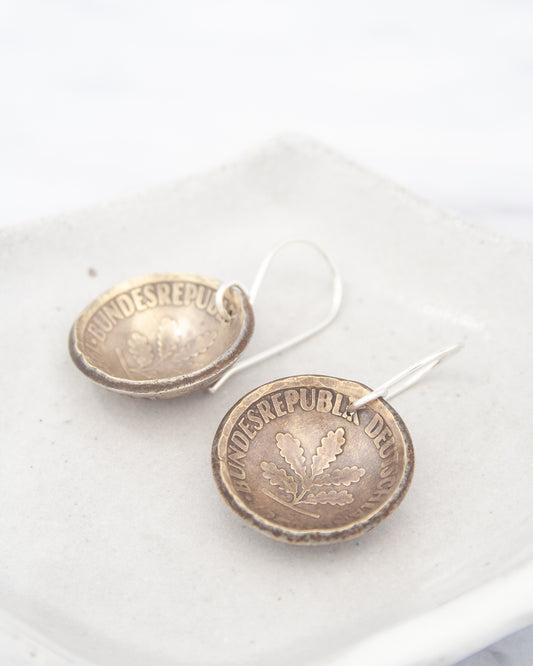 a pair of earrings made of a shiny metal, possibly silver, with the word "BUNDESREPULS" engraved on them. The earrings are placed on a white surface, such as a plate or a tray.