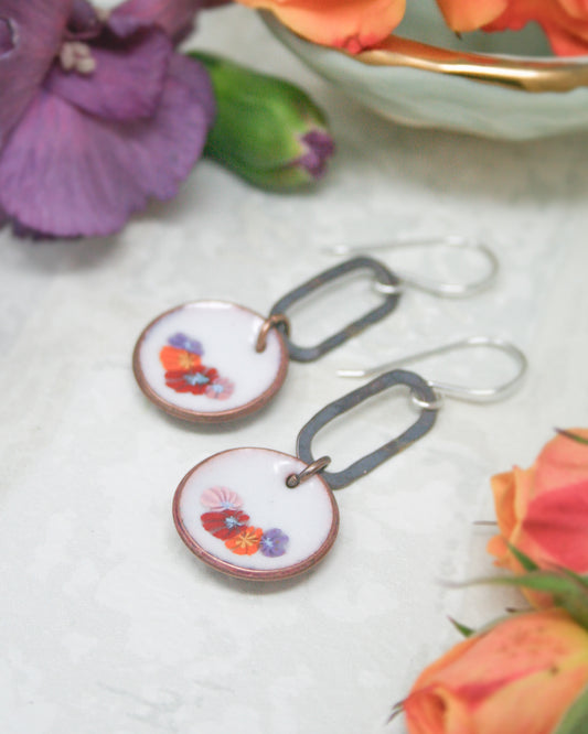 A pair of earrings with floral designs on the round pendants, placed on a white surface with a bowl of flowers and orange flowers in the background.