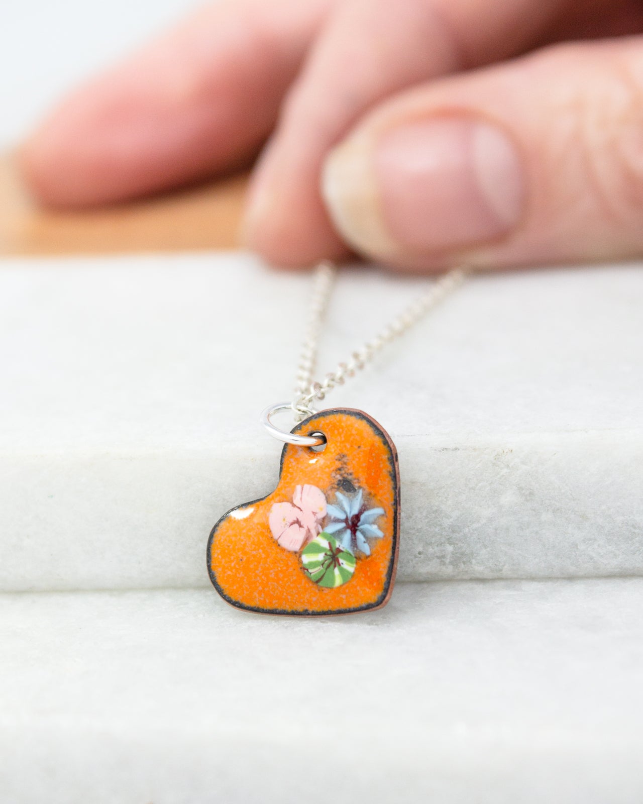 A hand is holding a heart-shaped pendant with a floral design, which is attached to a chain.