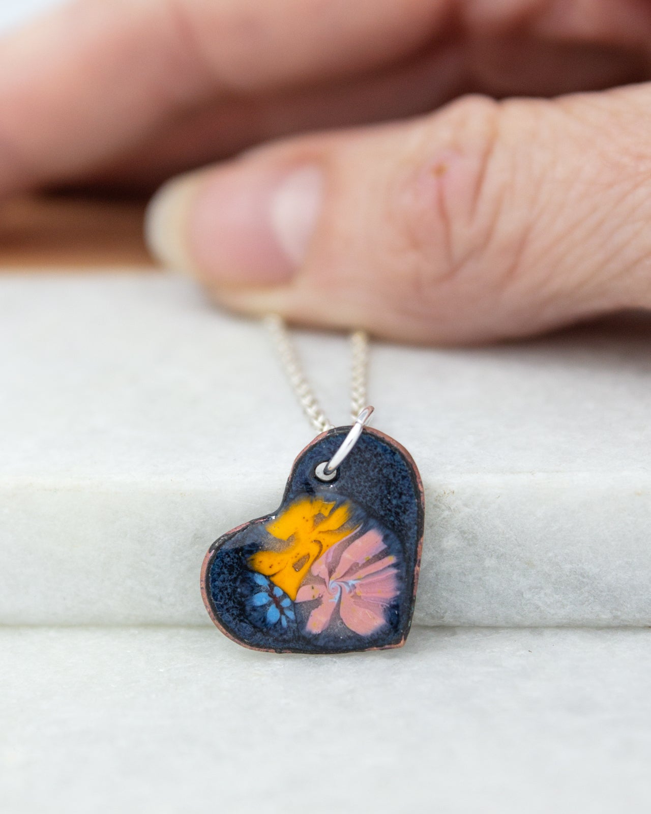 A hand is holding a heart-shaped pendant with a colorful floral design, resting on a white surface.