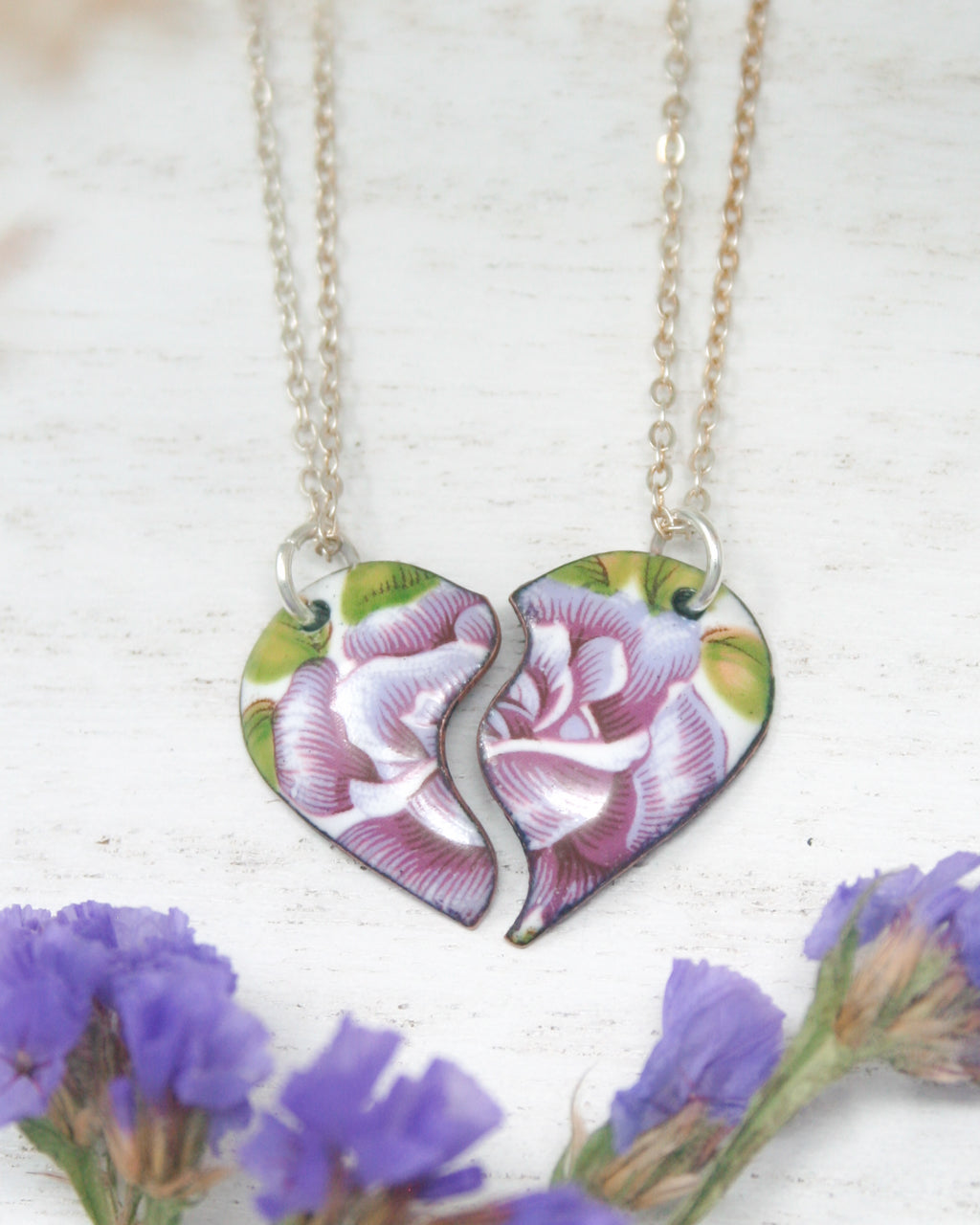 two broken heart shaped necklaces sitting next to purple flowers