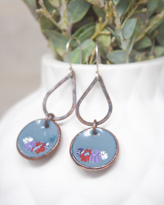 A pair of earrings with blue enamel and floral designs, hanging from a white vase with greenery.