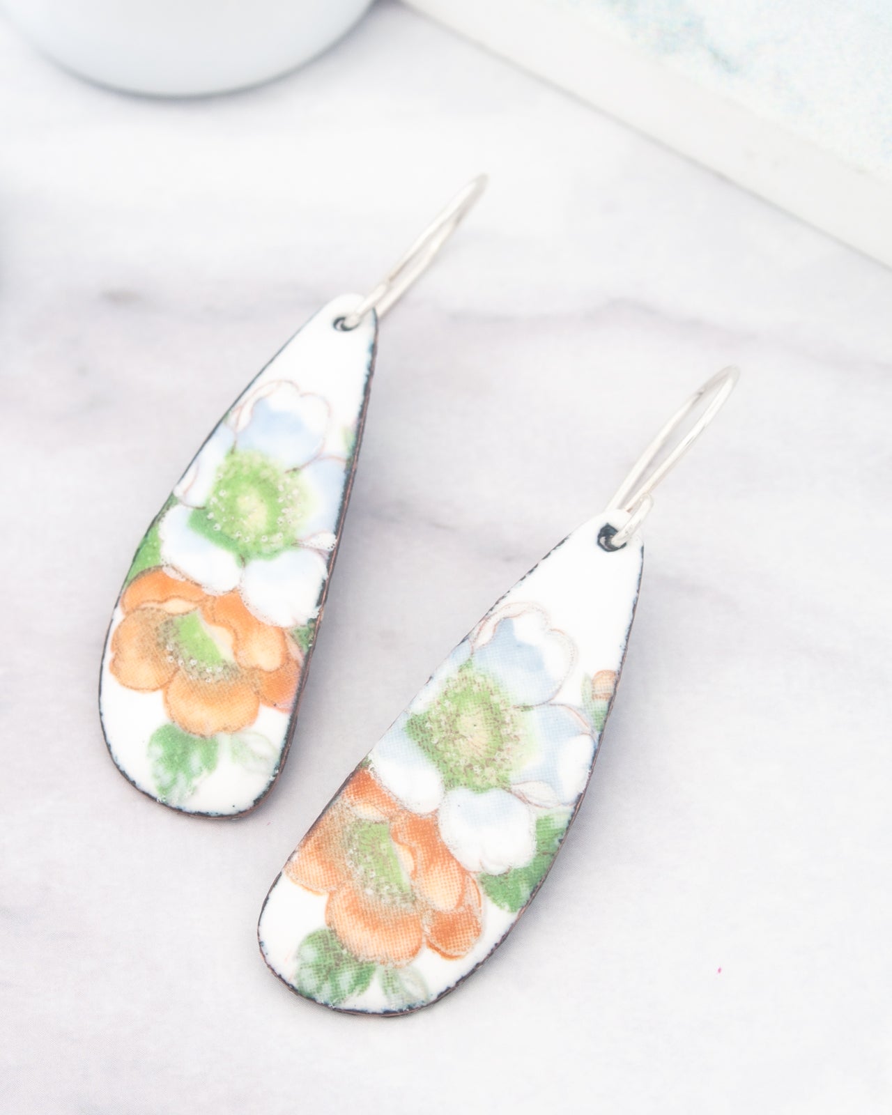 A pair of earrings with a floral design, featuring orange and green flowers, are displayed on a marble surface.