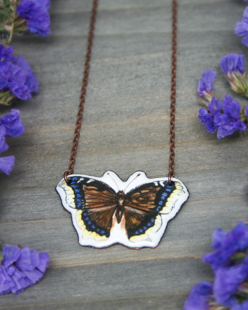 A necklace with a pendant in the shape of a butterfly is displayed on a wooden surface, surrounded by purple flowers.