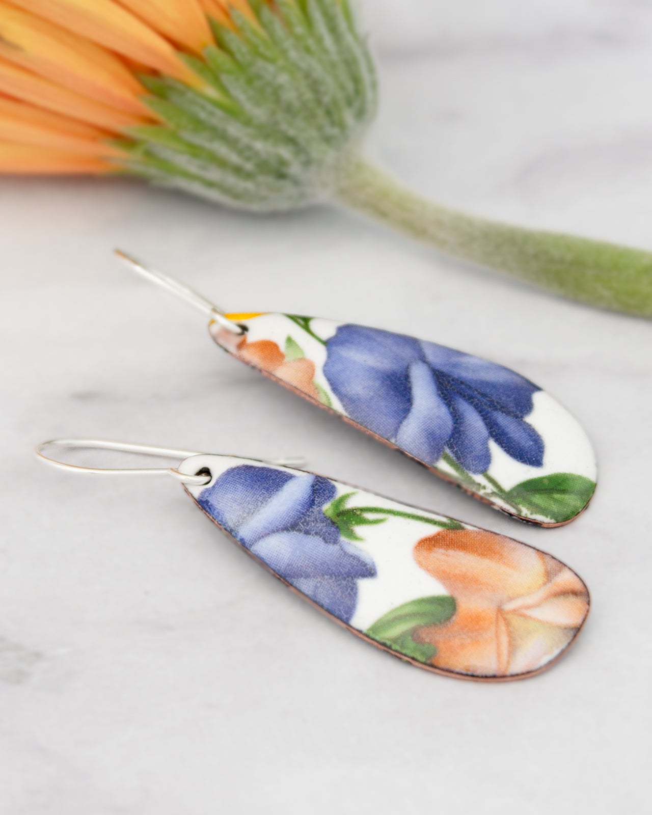 a pair of earrings with a floral design, including blue, orange, and white colors. The earrings are placed on a marble surface, with a flower in the background.