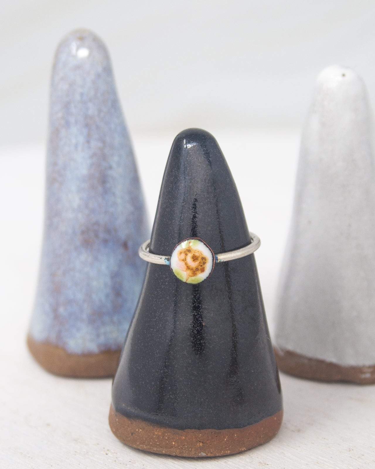 A silver ring with a flower design is displayed on a black ceramic stand, with two other ceramic stands in the background.