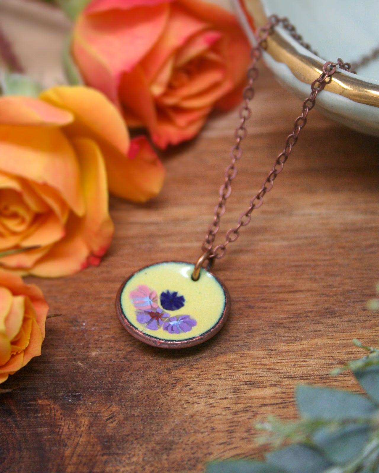 A necklace with a round pendant featuring a floral design rests on a wooden surface, surrounded by vibrant orange and yellow roses.