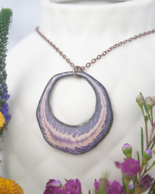 A necklace with a large, circular pendant featuring a purple and white design hangs from a chain. The necklace is displayed against a backdrop of colorful flowers, creating a visually appealing and vibrant scene.