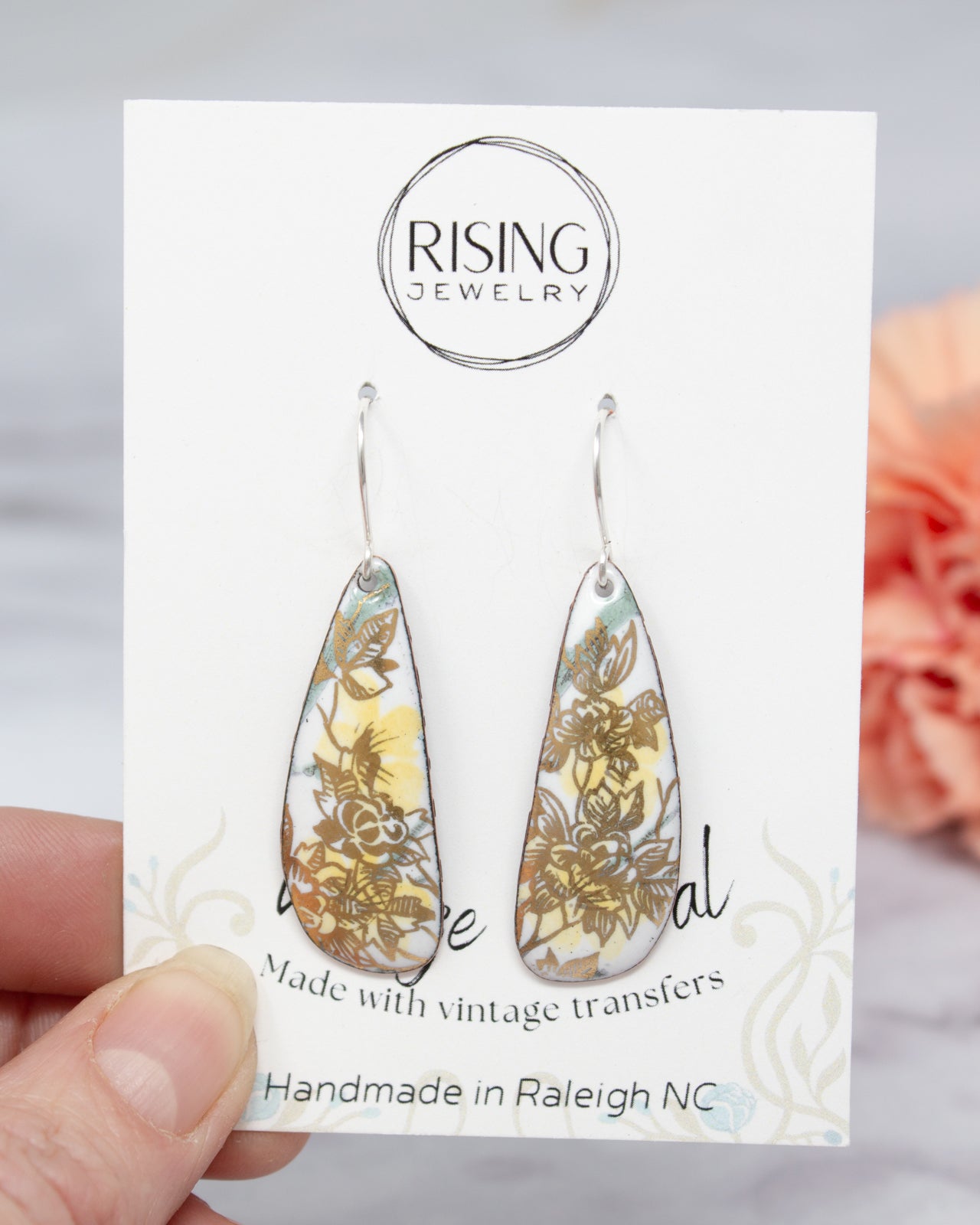 A hand holding a pair of earrings with a floral design, as shown on the packaging.
