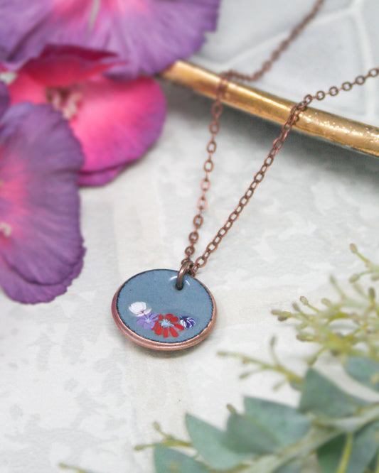 A necklace with a blue pendant featuring a floral design rests on a white surface, surrounded by purple and green flowers.