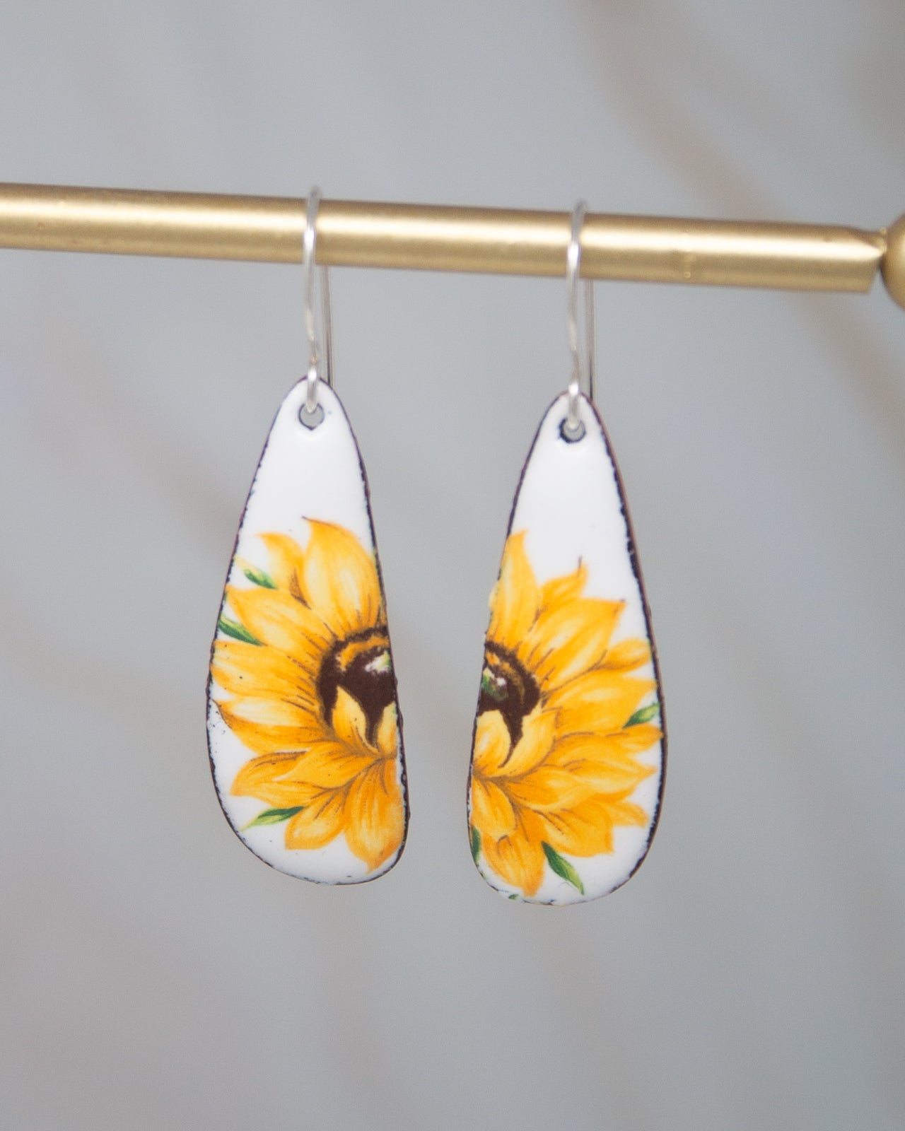 A pair of earrings with a sunflower design on them, hanging from a gold-colored metal hook.